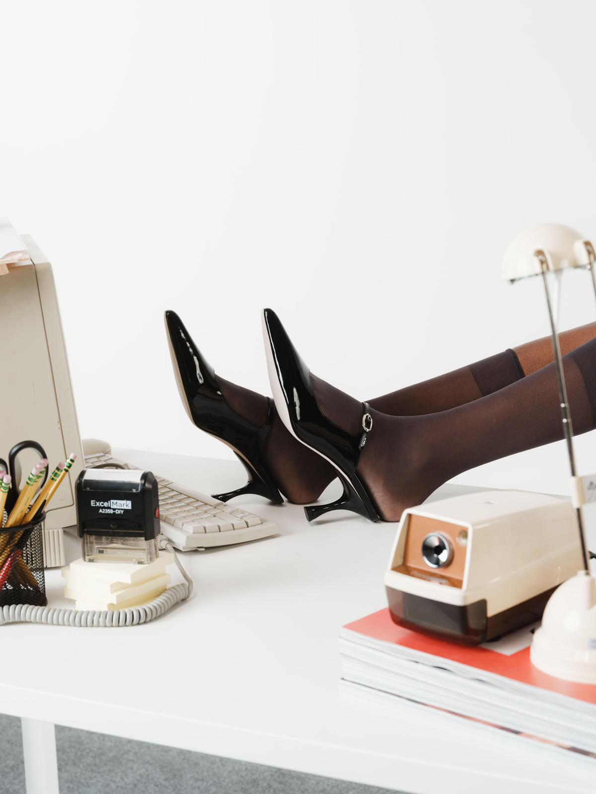 Editorial image of heels resting on a desk beside a keyboard and stamp